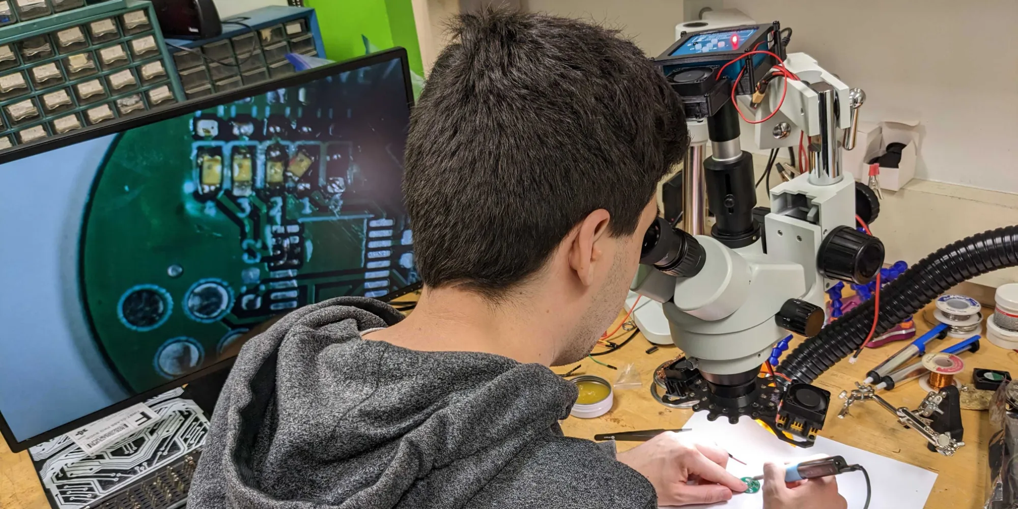 a man looks through a microscope to solder with a large screen on the left showing the circuit board being soldered